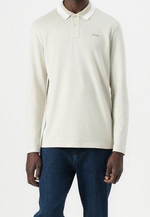 Mand iført en langærmet beige polo-shirt med knapper og mørkeblå jeans, står mod en ensfarvet baggrund.