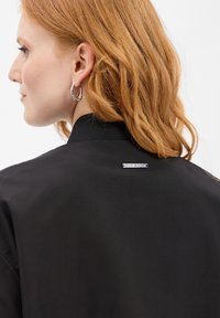 Czarna kurtka typu bomber z prążkowanym kołnierzem, gładką fakturą i srebrnym akcentem w postaci logo na plecach. Modelka ma na sobie duże kolczyki-koła.