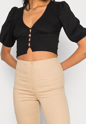 Vrouw draagt een zwarte cropped blouse met pofmouwen en knopen aan de voorkant, gecombineerd met beige broek met hoge taille, staand met handen in de zij.