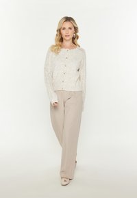 Cardigan lavorato a maglia color beige chiaro con scollo a zig zag, bottoni in legno e maniche a pizzo, abbinato a pantaloni a gamba larga beige chiaro.