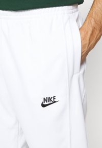 Vita atletiska byxor med elastisk midja och sidofickor, med en svart broderad Nike-logotyp på vänster ben.
