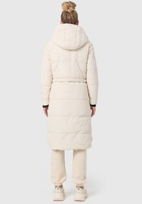 Lång, puffig, ljusbeige kappa med huva, bälte i midjan och vadderad design. Matchas med ljusbeiga byxor och vita sneakers.
