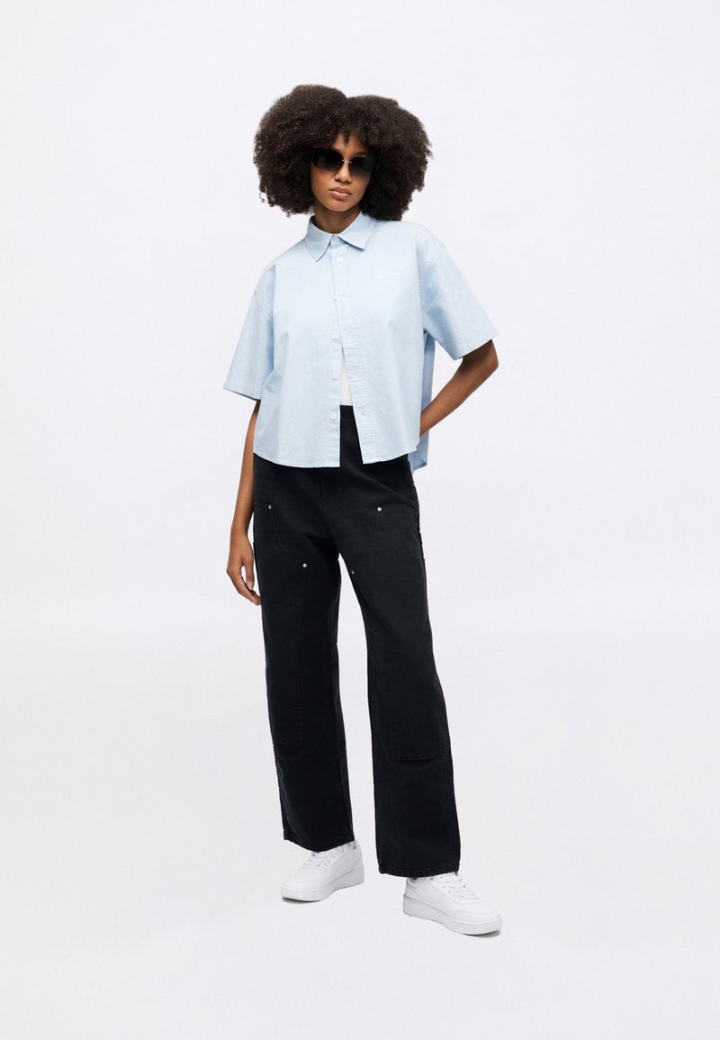 Chemise bleu clair courte à boutons, pantalon large noir avec des accents métalliques argentés, et baskets blanches ; modèle portant des lunettes de soleil foncées.