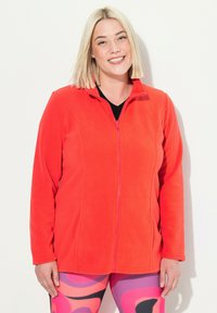 Punainen fleece-takki korkealla kauluksella ja täysivetoketjulla, jossa on pitkät hihat ja sileä pinta, yhdistettynä värikkäisiin kuvioituihin legginsseihin.