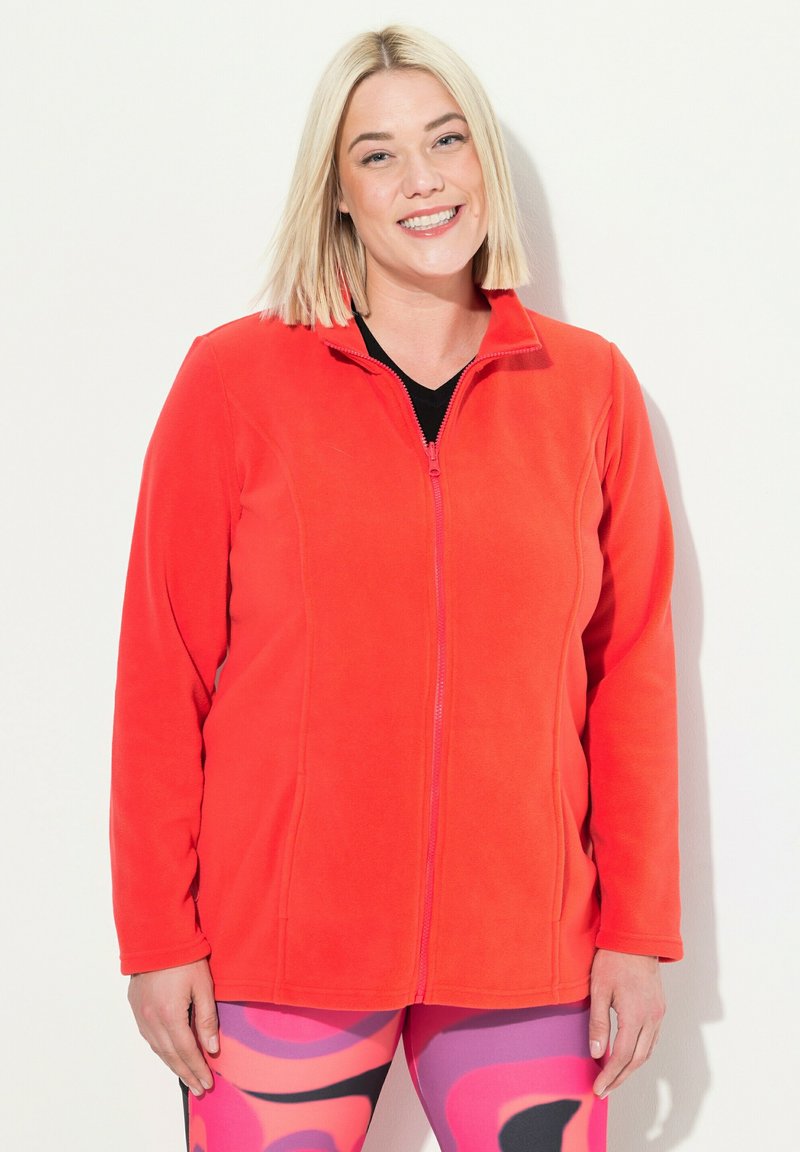 Giacca di pile rossa con colletto alto e zip completa, dotata di maniche lunghe e tessuto morbido, abbinata a leggings colorati con fantasie.