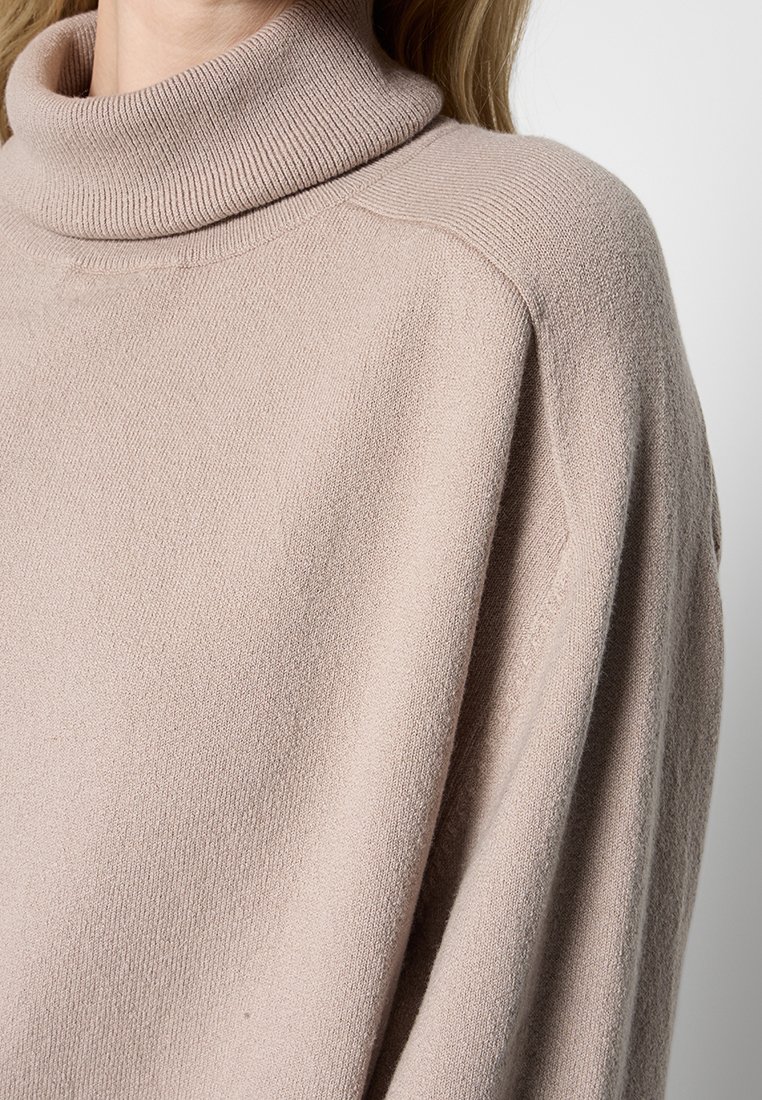 Beige Rollkragenpullover aus weichem Strickmaterial, mit weiter Passform und geripptem Kragen, sowie einer glatten, strukturierten Oberfläche.