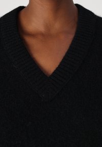 Pull noir en tricot avec un col en V profond. Présente une texture côtelée au niveau du col et un tissu doux et en peluche sur toute la surface.