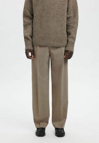 Brun strikket genser med ribbestrikkede mansjetter kombinert med beige bukser med vide ben og svarte slip-on sko, som viser en avslappet lagvis stil.