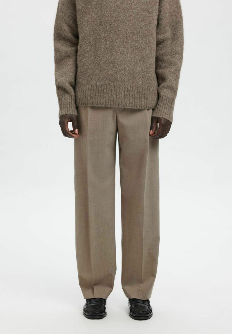 Brun strikket genser med ribbestrikkede mansjetter kombinert med beige bukser med vide ben og svarte slip-on sko, som viser en avslappet lagvis stil.