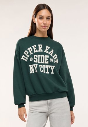 Kobieta z długimi ciemnymi włosami, ubrana w ciemnozieloną bluzę z białym napisem "Upper East Side NY City 1997" i jasnoszare dżinsy.