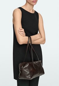 Femme portant une robe noire sans manches, tenant un sac à main en cuir marron foncé avec double poignée et fermeture éclair, sur un fond clair.
