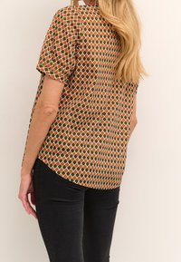 Blouse à manches courtes avec motif géométrique vert, orange et blanc. Tissu léger avec un ourlet arrondi, portée avec un pantalon noir ajusté.