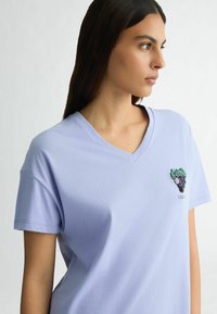 T-shirt lilas clair à col en V réalisé en tissu doux, avec un design de raisins perlés sur la poitrine gauche, à manches courtes et coupe décontractée.
