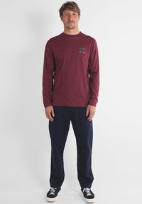 Chemise à manches longues bordeaux avec un motif subtil sur la poitrine, associée à un pantalon bleu marine et des baskets noires. Tissu lisse, coupe décontractée.