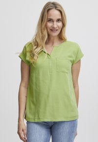 Maglietta verde a maniche corte con scollo a V, con tasca frontale e texture liscia. Abbinata a jeans blu chiaro.