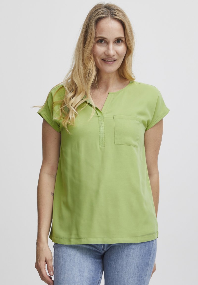 Maglietta verde a maniche corte con scollo a V, con tasca frontale e texture liscia. Abbinata a jeans blu chiaro.