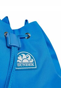Borsa a cordino blu in nylon con dettagli in ottone, con un patch a forma di cerchio in bianco e azzurro che raffigura delle onde.