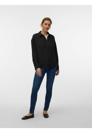 Kobieta stojąca, ubrana w czarną koszulę w cienkie prążki z guzikami, ciemnoniebieskie obcisłe dżinsy i czarne baleriny, na tle jasnego, jednolitego tła.