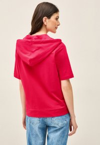 Red hooded sweatshirt with short sleeves, made of soft cotton. Simple design with a kangaroo pocket and relaxed fit, paired with light blue jeans.