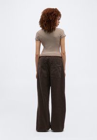 Γυναίκα με σγουρά μαλλιά, ντυμένη με εφαρμοστή καφέ και λευκή ριγέ κοντομάνικη μπλούζα και καφέ σκούρο παντελόνι με φαρδιά μπατζάκια, γυρισμένη προς τα πίσω.