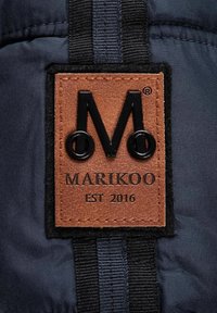 Bruin leren label met een reliëf zwart logo "M", "MARIKOO" en "EST 2016", gestikt op een gestructureerde donkere stoffen achtergrond.