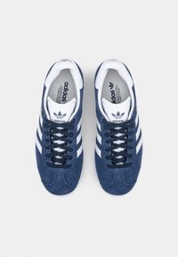 Modré semišové sportovní boty s bílými pruhy, bílým polstrovaným jazykem a tmavě modrými tkaničkami. Na straně a stélce je logo Adidas.