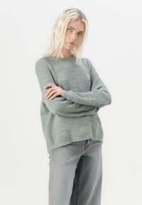 Lys grønn strikket genser med avslappet passform, teksturert mønster og ribbet halsåpning. Stylert med grå jeans som har en litt lengre bakdel.