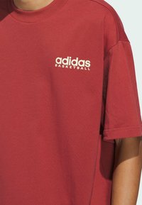 Červené bavlněné tričko s krátkými rukávy a kulatým výstřihem, na levé části hrudi je bílé text "adidas BASKETBALL".