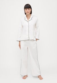 Set de pijama alb cu detalii negre; include o cămașă cu mânecă lungă și nasturi, precum și pantaloni lejeri. Material neted și design confortabil.