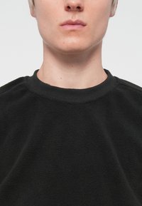 Svart fleecehäftad sweatshirt med ribbad krage, strukturerat tyg och sänkt axeldesign. Inga synliga logotyper eller mönster.
