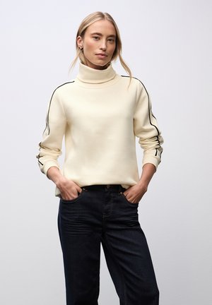 Frau in einem cremefarbenen Rollkragenpullover mit schwarzen Streifen auf den Ärmeln und dunkelblauen Jeans, steht mit den Händen in den Taschen.