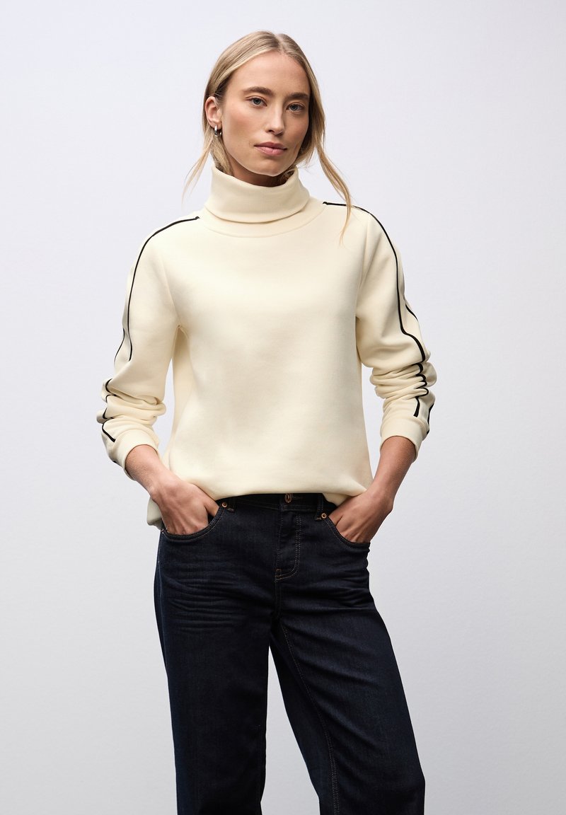Woman wearing a cream turtleneck sweater with black stripes on the sleeves and dark blue jeans, standing with hands in pockets.