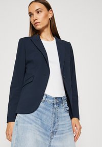 Marineblazer mit taillierter Passform, Ein-Knopf-Verschluss und zwei Fronttaschen. Kombiniert mit einem gerippten weißen Hemd und einem hellen Jeansrock.