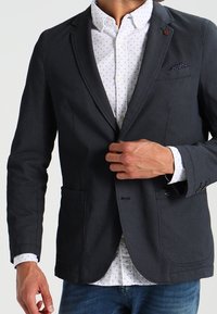 Dunkelgrauer Blazer aus strukturiertem Stoff, mit Ein-Knopf-Verschluss, zwei Fronttaschen und einem Einstecktuch, auf einem weißen gemusterten Hemd getragen.