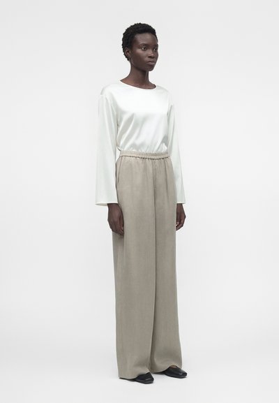 Γυναίκα που στέκεται, φορώντας λευκό σατινέ μπλουζάκι με μακριά μανίκια, μπεζ παντελόνια με φαρδιά μπατζάκια και ελαστική μέση, και μαύρα μυτερά παπούτσια σε απλό φόντο.