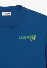 Camiseta de algodón azul de cuello redondo con el logo verde de "Lacoste" y un cocodrilo bordado. Textura suave, corte clásico y mangas cortas.