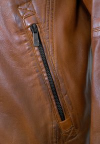 Braune Lederjacke mit einem schwarzen Reißverschluss, strukturierten Oberflächen und kontrastierenden Nähten. Der Reißverschluss befindet sich auf der linken Seite.