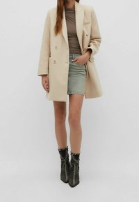 Beige double-breasted coat, fitted brown top, light denim skirt, and textured black ankle boots with a folded cuff, standing against a gray background.