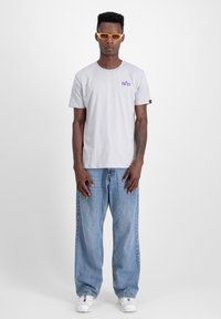 Hall grey puuvillane T-särk, millel on lilla graafiline logo vasakus rinnas, koos heledad sinised vabalt lõigatud teksade ja valgete tossudega.