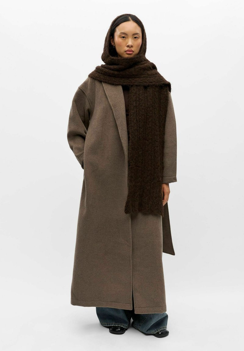 Femme portant un long manteau marron, une écharpe marron foncé oversize enroulée autour du cou et de la tête, un jean large et des chaussures noires, debout les mains dans les poches.
