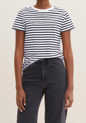 Person iført en hvid og sort vandretstribet kortærmet skjorte, stoppet ned i højtaljede sorte jeans, stående med armene afslappet ned langs siden.