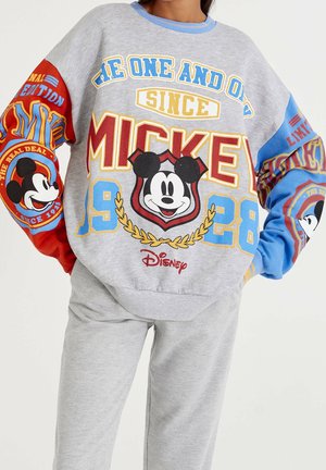 Osoba nosząca szarą bluzę i spodnie z kolorowymi grafikami Myszki Miki oraz tekstem odnoszącym się do "Mickey 1928" i marki Disney.