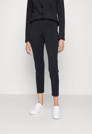 Persoon draagt strakke zwarte broek, zwarte knoopjasje en witte sneakers tegen een effen lichte achtergrond.