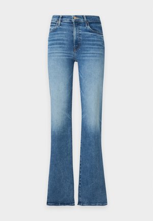 Femme debout portant un haut gris clair à manches longues, un jean droit taille haute bleu et des chaussures blanches à talons pointus sur fond blanc.