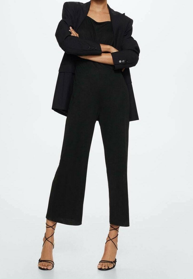 Personne portant un blazer noir par-dessus une combinaison noire sans manches à jambes larges et des sandales noires à talons hauts avec des lanières, debout bras croisés.