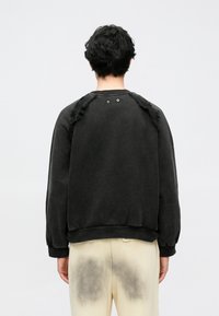 Sweatshirt preta com detalhes texturizados e desfiados nos ombros e botões de pressão metálicos na parte de trás. Punhos e bainha canelados. Tecido macio e mate.