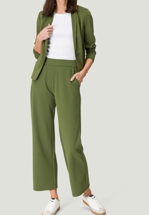 Femme portant un blazer vert olive et un pantalon large assorti avec un haut blanc et des baskets blanches, debout devant un fond uni.