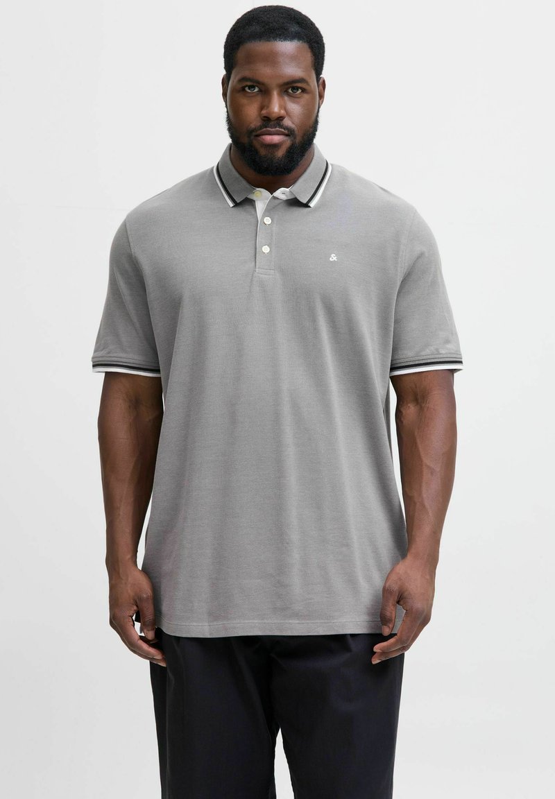 Man met een baard in een lichtgrijze polo met een zwart-witte gestreepte kraag en mouwen, staande tegen een effen witte achtergrond.