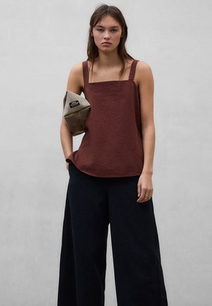 Kobieta ubrana w brązowy, bezrękawnikowy top o kwadratowym dekolcie i luźne czarne spodnie, trzymająca pod pachą składany beżowo-szary kopertówkę.