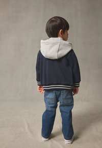 Giacca blu navy con cappuccio grigio, polsini a costine e strisce bianche. Jeans in denim azzurro chiaro con due tasche posteriori e scarpe da ginnastica bianche.